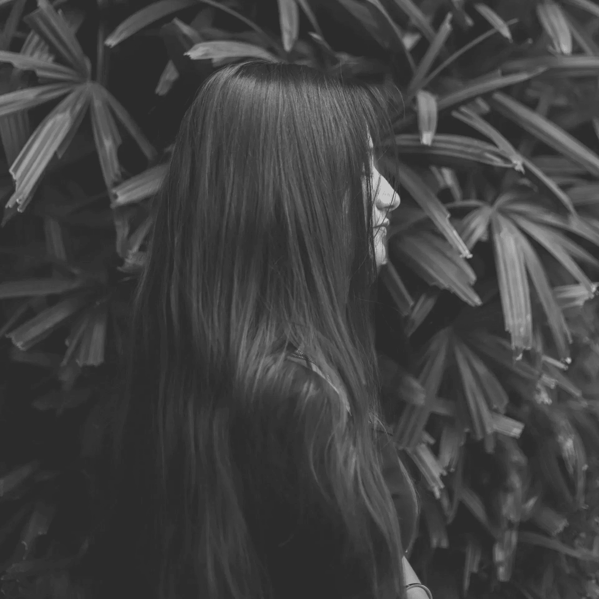 A woman with long hair stands in front of lush green tropical plants.