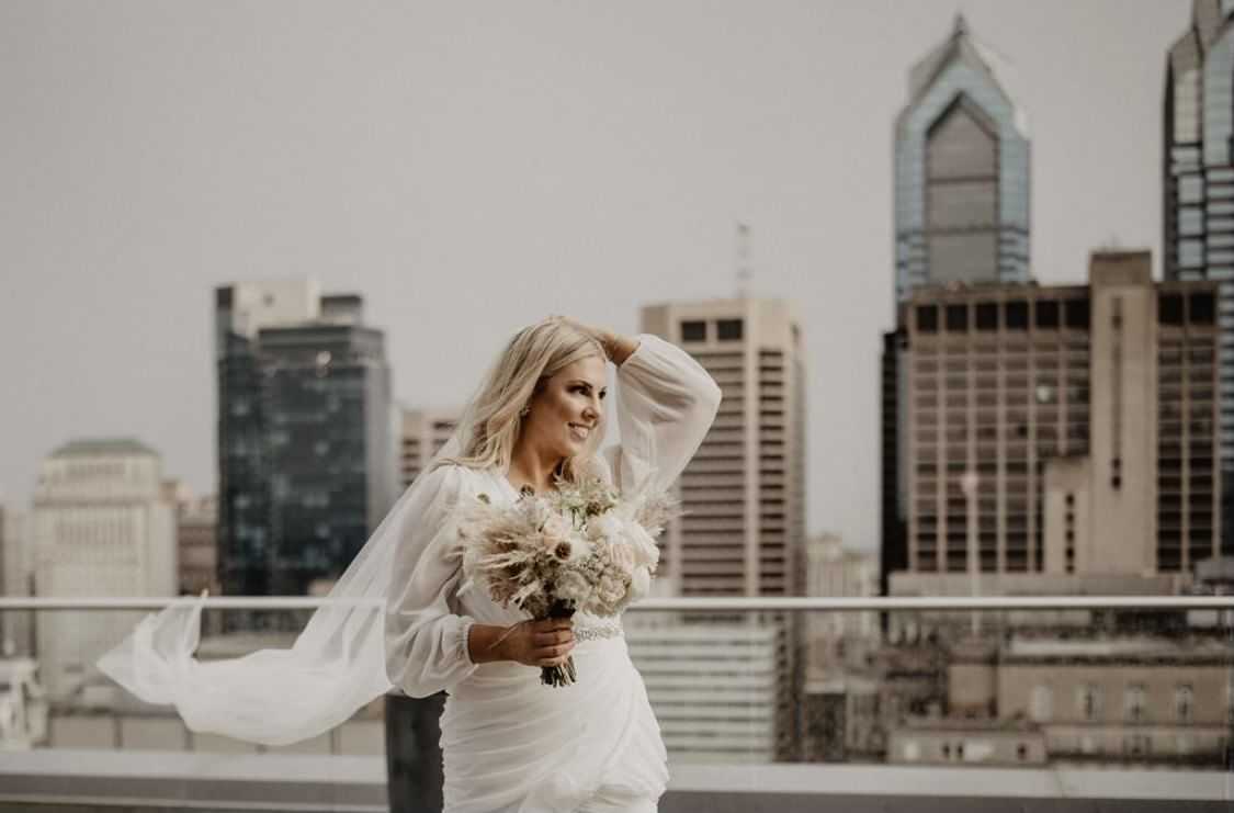 Bride with bouquet smiles on rooftop with city skyline in background.